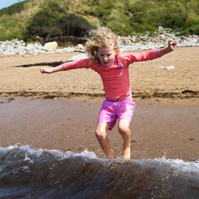 A child in a UV Protective Rash Vest Red leaps over a wave on a sandy beach, arms outstretched, hair blowing in the wind, under a sunny sky.